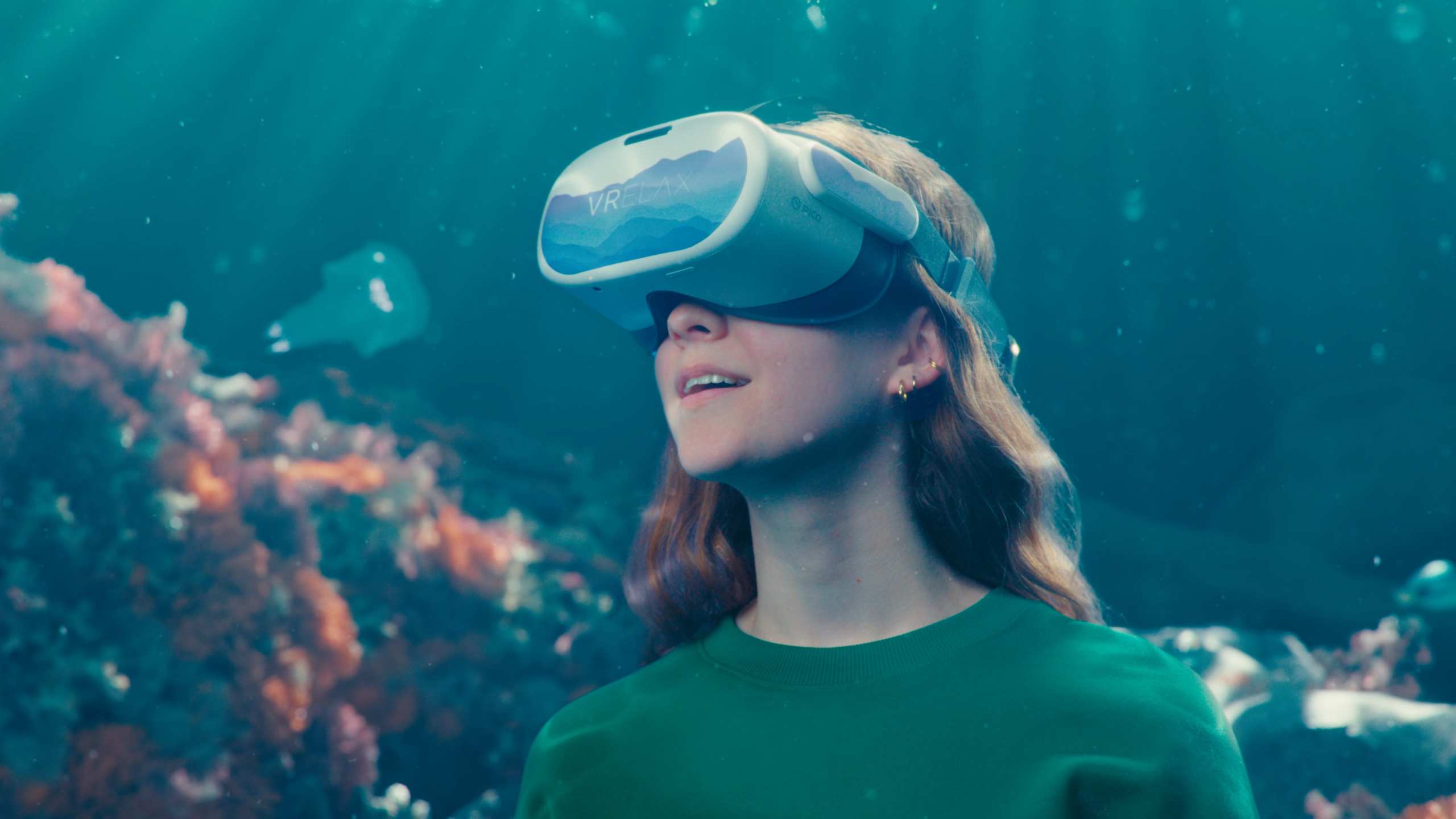 A smiling young woman wearing a virtual reality (VR) headset and surrounded by a virtual underwater environment, taken from Sensu.health's commercial video for VRelax