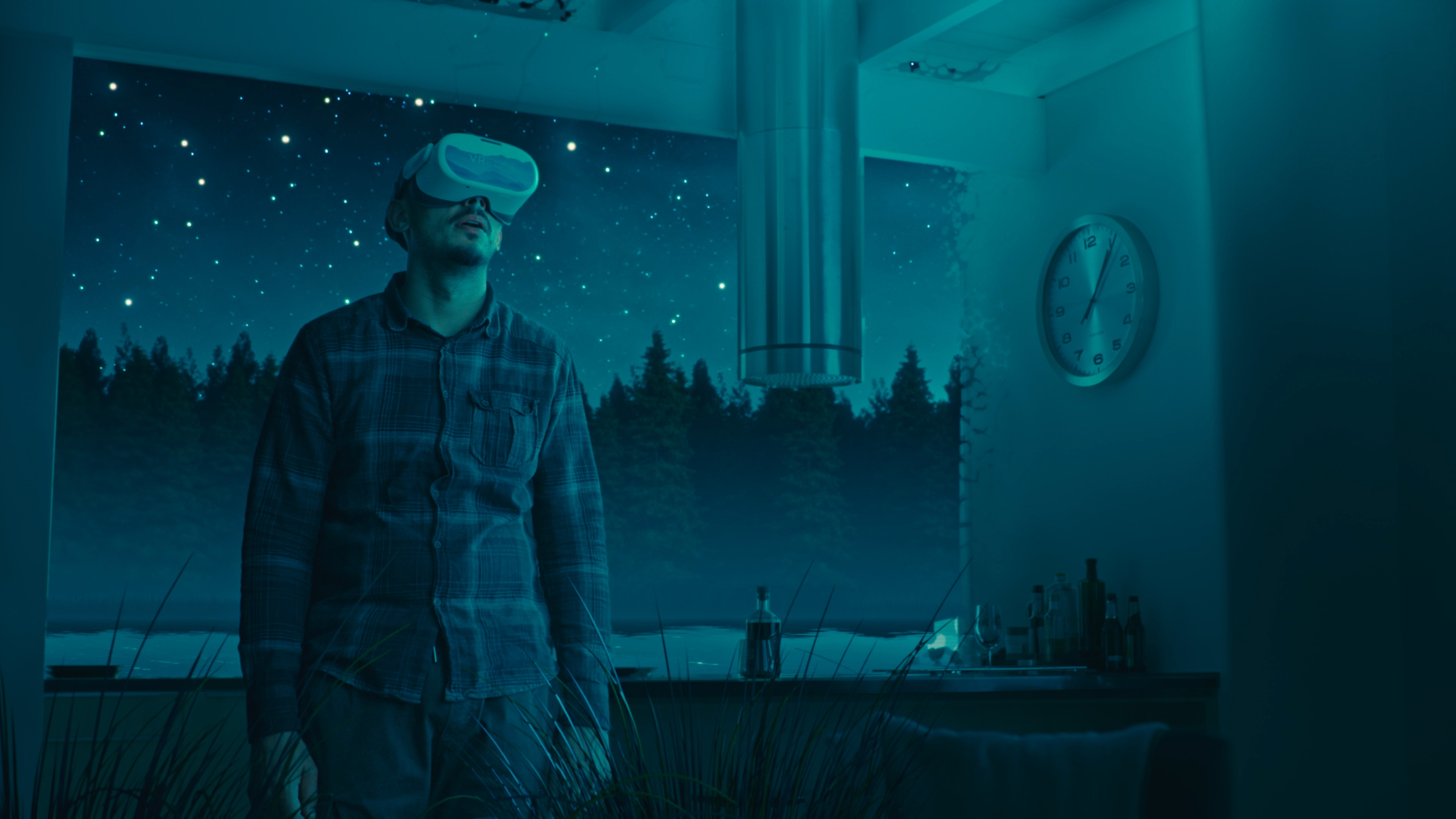 A man wearing a virtual reality (VR) headset is seen partly in domestic surroundings and partly in a virtual outdoor environment, with a backdrop of woodlands and a night sky full of stars (taken from Sensu.health's commercial video for VRelax)